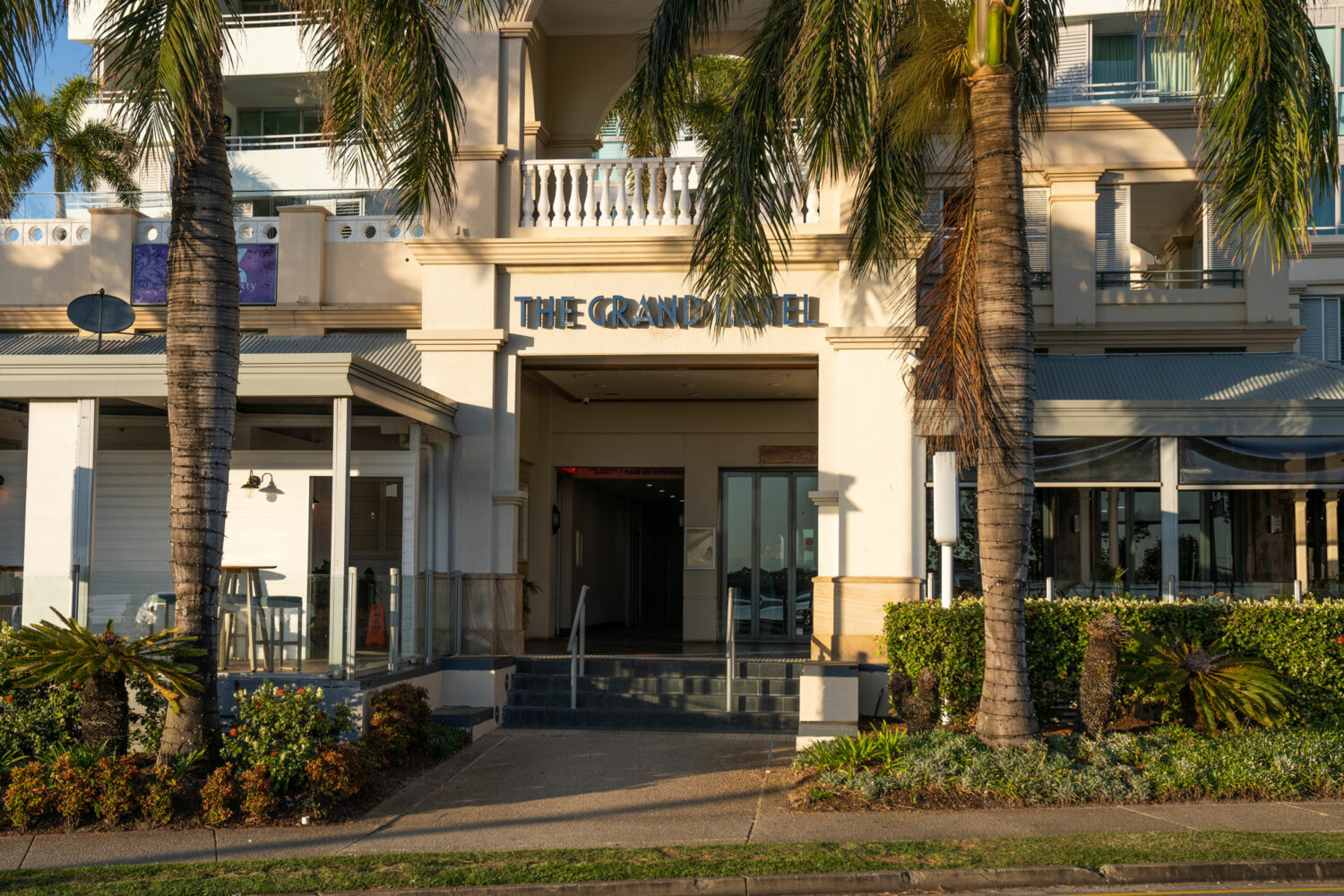 The entryway to "The Grand Hotel" building with large square columns and sign above the entrance within the Labrador lifestyle precinct. There are palm trees in the foreground and a few short steps from the footpath up to the door. It looks like an inviting, tropical oasis that could be the perfect place to retire.
