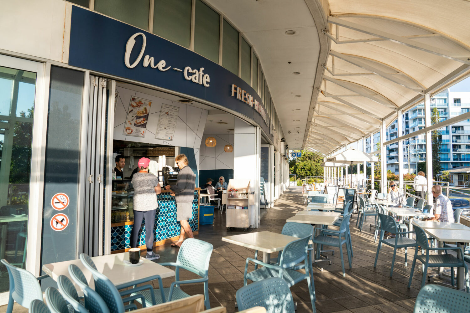 One Cafe at the Labrador PARK SHORE lifestyle village. It has a blue sign above the entrance with white writing. There are people placing an order for coffee at the counter.